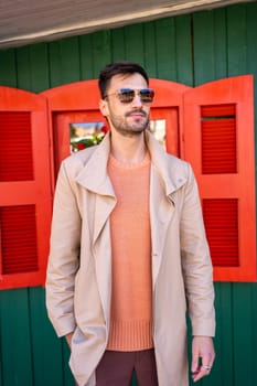 Fashionable man in sunglasses. Stylish and confident male wearing shades, salmon-color sweater and beige trench coat. Handsome guy with dark hair standing outside Christmas house.