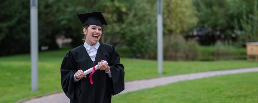 Portrait of happy caucasian woman in graduate gown holding diploma outdoors