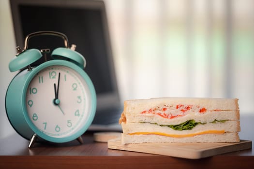 Sandwich and alarm clock displaying noon time on wooden table in office for the concept of fast food eating and time management at the workplace