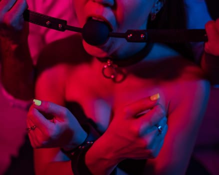 A man gags a woman in a pink blue neon light in the bedroom. BDSM concept