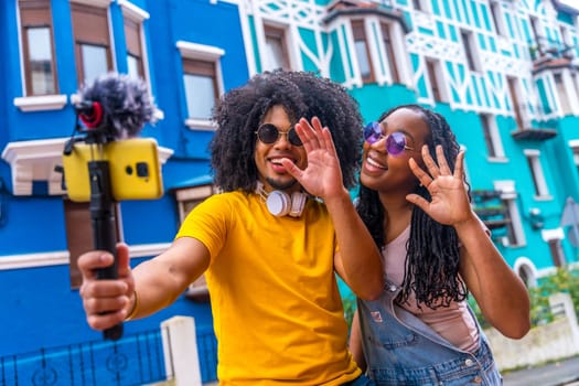 African couple broadcasting a life video waving at camera next to blue colorful house