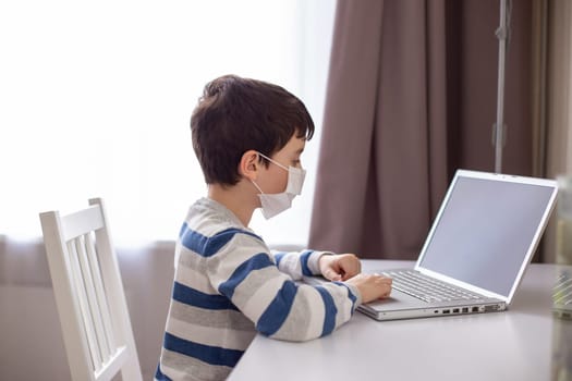 A boy in a striped T-shirt and a white medical mask, sits behind a monitor with a laptop in room , at home, distance learning during.