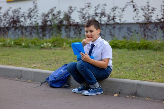schoolboy in a white shirt with a blue tie, holds a blue lunch box and eats a piece of apple, looks at the camera.