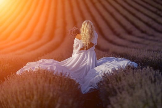 Blonde woman poses in lavender field at sunset. Happy woman in white dress holds lavender bouquet. Aromatherapy concept, lavender oil, photo session in lavender.