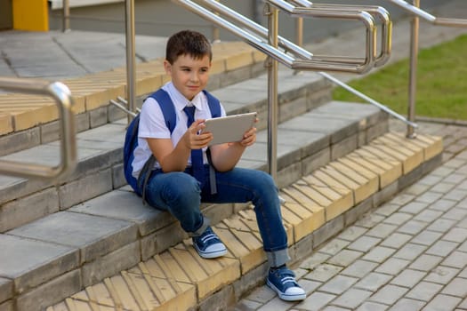 A beautiful schoolboy in a white shirt with a blue backpack, blue tie, blue jeans sits on the stairs outside and holds with a gray tablet, looks to the side.