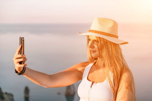 Selfie woman in a hat, white tank top, and shorts captures a selfie shot with her mobile phone against the backdrop of a serene beach and blue sea