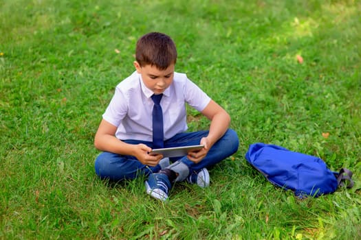Serious Schoolboy in a white shirt and blue tie sits on green grass against a blue sky with white clouds and looks into a tablet. Copy space. Top view