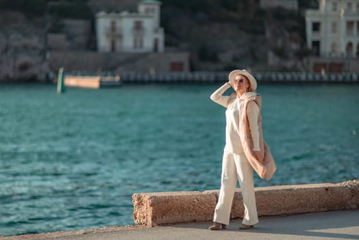 Happy blonde woman in a white suit and hat posing at the camera against the backdrop of the sea.