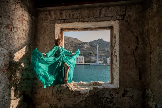 Rear view of a happy blonde woman in a long mint dress posing against the backdrop of the sea in an old building with columns. Girl in nature against the blue sky