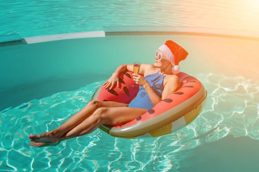 A happy woman in a blue bikini, a red and white Santa hat and sunglasses poses in the pool in an inflatable circle with a watermelon pattern, holding a glass of champagne in her hands. Christmas holidays concept