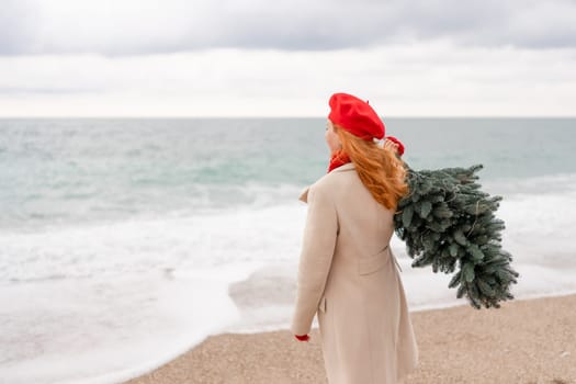 Redhead woman Christmas tree sea. Christmas portrait of a happy redhead woman walking along the beach and holding a Christmas tree on her shoulder. She is dressed in a light coat and a red beret