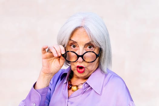 portrait of a friendly surprised senior woman looking at camera over her glasses, concept of elderly people happiness and active lifestyle, copy space for text