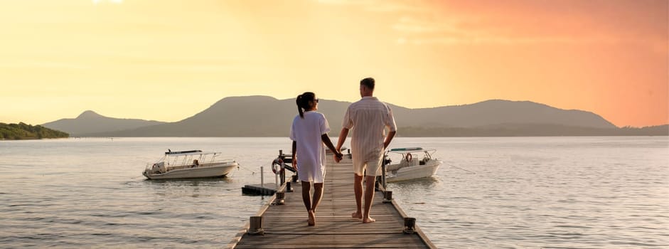 couple walking on a wooden pier in the ocean at sunset in Thailand.Caucasian men and Asian women diverse couple waking at a jetty in the ocean, man and women watching sunset together