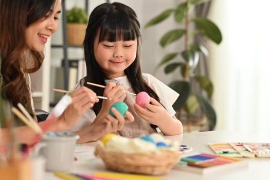 Cute little child girl painting Easter eggs with her mother in living room.