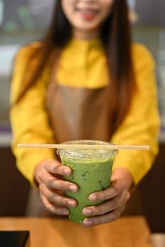 Smiling female cafe owner holding a plastic cup of iced matcha green tea latte with paper straw.