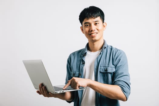 Confidence shines as a young Asian man smiles while working on a laptop, sending emails or chatting online. Studio shot isolated on white background, capturing his enthusiasm for digital communication