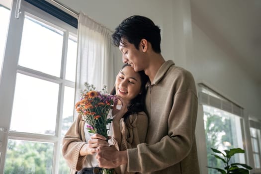 Romantic young asian couple embracing with holding flowers and smiling in living room at home. fall in love. Valentine concept.