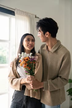 Romantic young asian couple embracing with holding flowers and smiling in living room at home. fall in love. Valentine concept.