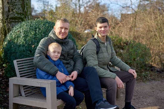 Dad with two sons sitting on a bench in autumn park. Experience the tranquility of familial bonds in the heart of autumn with this serene image. A father, 40 years old, and his two sons - a beautiful 8-year-old boy and a 17-year-old young man, seated in the park. The autumnal ambiance adds warmth to this captivating moment of family togetherness.