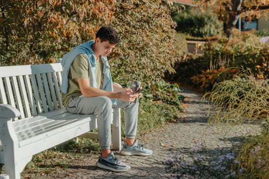 a teenager sits on a bench in the park drinks coffee from a thermo mug and looks into a phone. Portrait of handsome cheerful guy sitting on bench fresh air using device browsing media smm drinking latte urban outside outdoor.