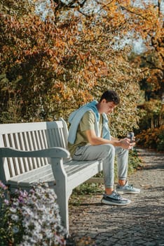 a teenager sits on a bench in the park drinks coffee from a thermo mug and looks into a phone. Portrait of handsome cheerful guy sitting on bench fresh air using device browsing media smm drinking latte urban outside outdoor.