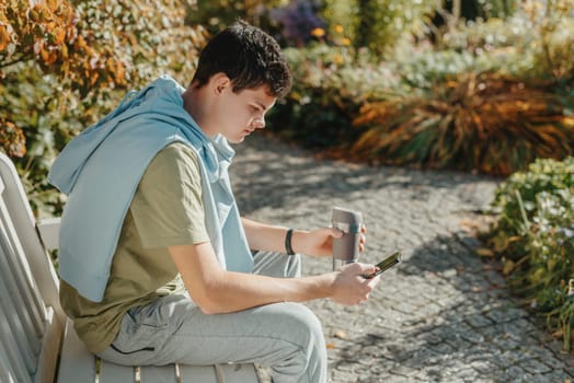 a teenager sits on a bench in the park drinks coffee from a thermo mug and looks into a phone. Portrait of handsome cheerful guy sitting on bench fresh air using device browsing media smm drinking latte urban outside outdoor.