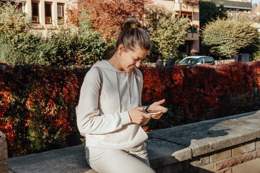 Young fashionable teenage girl with smartphone in park in autumn sitting at smiling. Trendy young woman in fall in park texting. Retouched, vibrant colors. Beautiful blonde teenage girl wearing casual modern autumn outfit sitting in park in autumn. Retouched, vibrant colors, brownish tones.