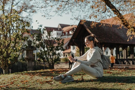 Young fashionable teenage girl with smartphone in park in autumn sitting at smiling. Trendy young woman in fall in park texting. Retouched, vibrant colors. Beautiful blonde teenage girl wearing casual modern autumn outfit sitting in park in autumn. Retouched, vibrant colors, brownish tones.