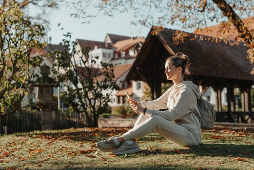 Young fashionable teenage girl with smartphone in park in autumn sitting at smiling. Trendy young woman in fall in park texting. Retouched, vibrant colors. Beautiful blonde teenage girl wearing casual modern autumn outfit sitting in park in autumn. Retouched, vibrant colors, brownish tones.