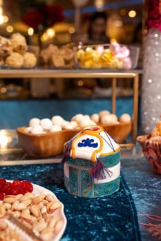 Yurt-shaped container with intricate patterns sits on a blue tablecloth, surrounded by an assortment of sweets and nuts. The background features a blurred buffet setting.