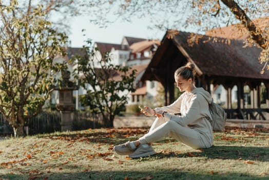 Young fashionable teenage girl with smartphone in park in autumn sitting at smiling. Trendy young woman in fall in park texting. Retouched, vibrant colors. Beautiful blonde teenage girl wearing casual modern autumn outfit sitting in park in autumn. Retouched, vibrant colors, brownish tones.