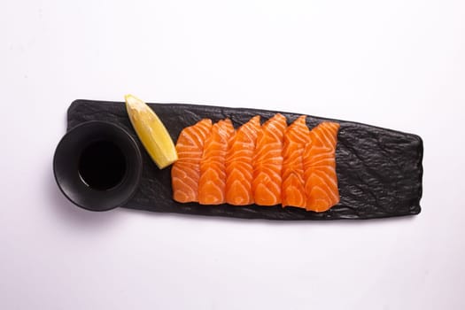 Top view of a delicious and healthy meal of fresh salmon fillet sliced into thin strips and served on a black stone plate with soy sauce and a lemon wedge. The plate is isolated on a white background.