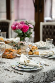 An elegant table setting with pink roses centerpiece in a crystal vase, fine china plates, silverware, and crystal wine glasses. White tablecloth, blurred background with window and light.