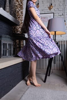 A young girl with long dark hair is dancing happily near the fireplace. She is wearing a purple dress with a white pattern and is barefoot. The girl is smiling and full of joy.