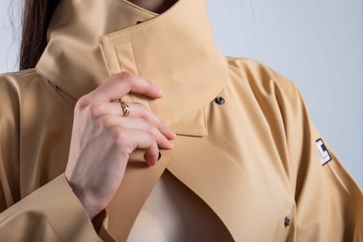 Elegant woman with nude makeup and pink lips in a stylish beige raincoat with black buttons, exuding sophistication and chicness. She poses near a white wall, showcasing a fashion-forward look.