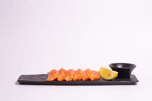 Top view of a delicious and healthy meal of fresh salmon fillet sliced into thin strips and served on a black stone plate with soy sauce and a lemon wedge. The plate is isolated on a white background.