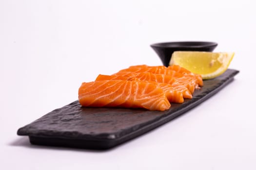 A close-up of a fresh salmon fillet garnished with lemon wedges and soy sauce on a black stone plate. This appetizing seafood dish is isolated on a white background, perfect for a healthy meal.