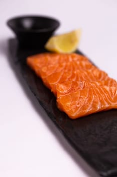 A close-up of a fresh salmon fillet garnished with lemon wedges and soy sauce on a black stone plate. This appetizing seafood dish is isolated on a white background, perfect for a healthy meal.