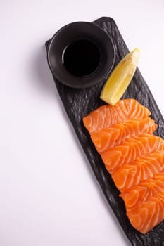 A close-up of a fresh salmon fillet garnished with lemon wedges and soy sauce on a black stone plate. This appetizing seafood dish is isolated on a white background, perfect for a healthy meal.