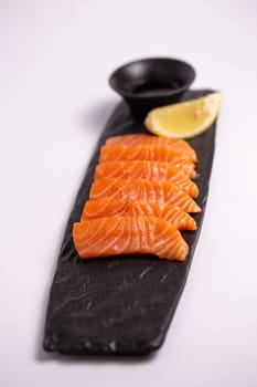 Top view of a delicious and healthy meal of fresh salmon fillet sliced into thin strips and served on a black stone plate with soy sauce and a lemon wedge. The plate is isolated on a white background.