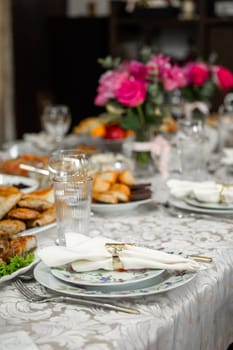 An elegant table setting with pink roses centerpiece in a crystal vase, fine china plates, silverware, and crystal wine glasses. White tablecloth, blurred background with window and light.