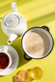 A cozy breakfast setup featuring oatmeal with milk, tea in a white cup on saucer, honey in a glass bowl, and bread slices on a yellow background. Shadows add depth and warmth to the scene.