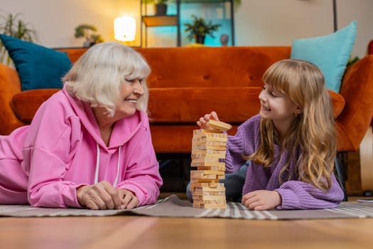 Excited little cute girl granddaughter playing building block game with smiling elderly grandparent grandmother. Building wooden tower and taking out blocks in living room at home during weekends.