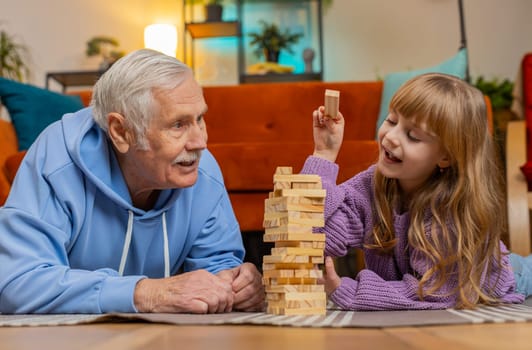 Excited little cute child girl granddaughter kid playing building block game with smiling elderly grandfather. Building wooden tower and taking out blocks in living room at home floor during weekends.
