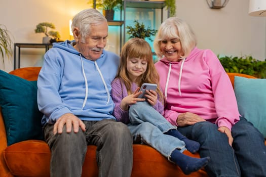 Cute Caucasian girl teaching grandfather and grandmother using smartphone on sofa in living room. Grandparents watching granddaughter play games on mobile phone. Happy family at home during weekend.