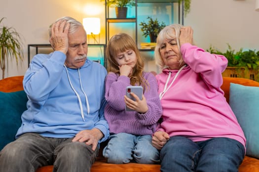 Upset grandfather, grandmother and granddaughter playing game on smartphone and losing at home. Sad Caucasian girl with grandparents received bad news, fortune loss, fail education study test on couch