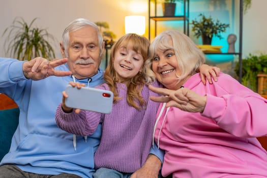 Happy grandfather, grandmother and granddaughter taking selfie photo with smartphone at home. Smiling Caucasian girl with grandparents make blogger self portrait showing peace sign sitting on couch