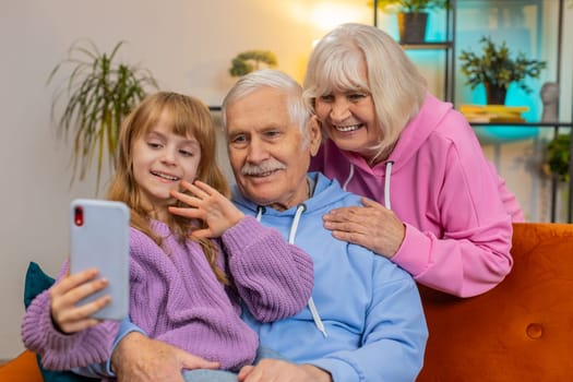 Caucasian grandfather, grandmother and small granddaughter making video call online on smartphone at home. Girl with senior grandparents talking by internet webcam chat app sitting on sofa in room.