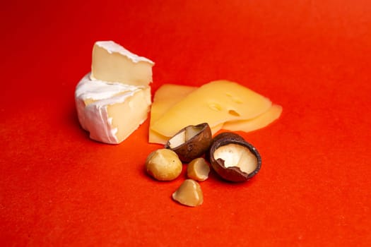 An exquisite assortment of brie, cheddar, and gouda cheeses elegantly presented on a vibrant red background, accompanied by a scattering of delicious macadamia nuts.