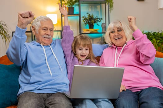 Surprise Caucasian grandfather, grandmother and granddaughter using laptop netbook while celebrating success at home. Happy girl with grandparents amazed by online game win clenching fists on couch